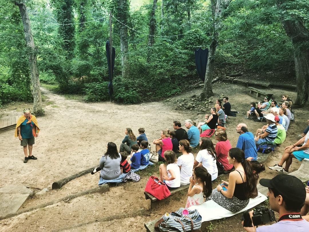 Families and theater lovers gather in a shaded forest clearing to enjoy an outdoor Shakespeare play. The natural surroundings and casual seating create a magical open-air performance in Tisbury.