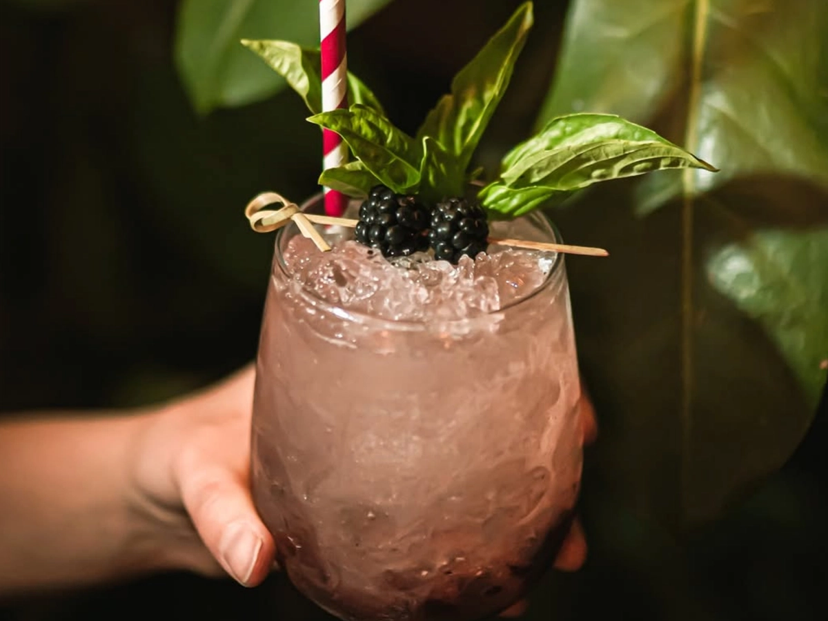 A hand holding a cocktail glass filled with crushed ice, garnished with fresh blackberries, basil leaves, and a red-and-white striped straw, set against a backdrop of lush green leaves.