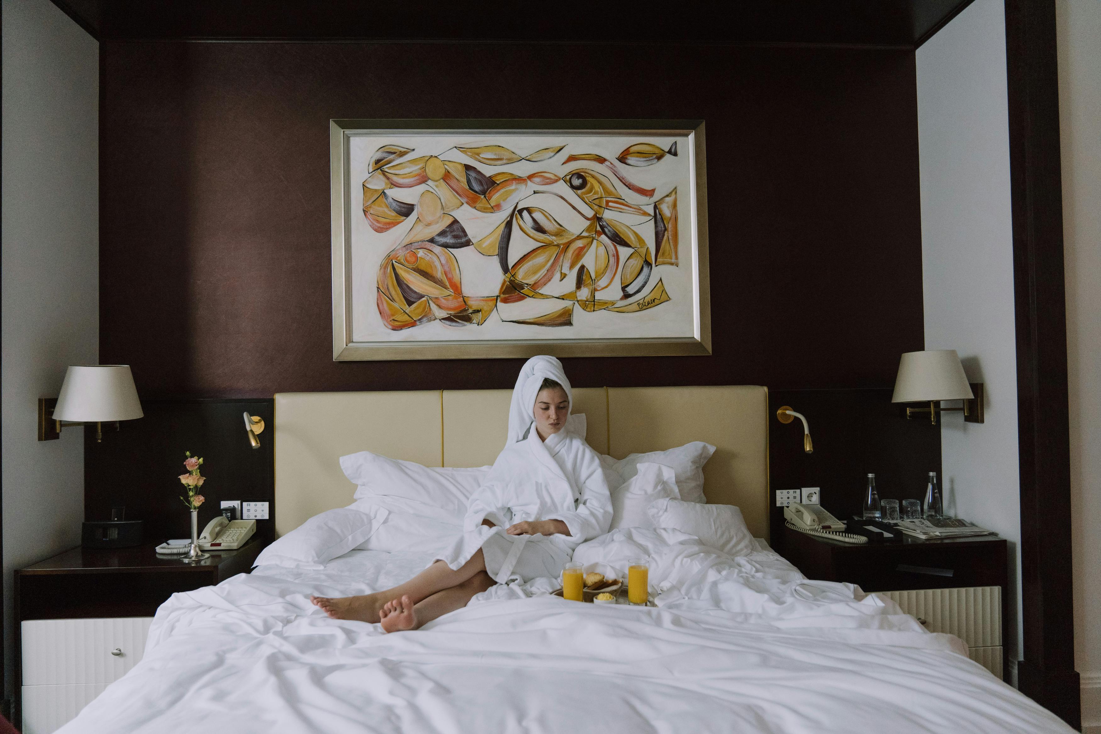 A woman relaxes in a plush hotel bed wearing a robe and towel wrap, with breakfast served on a tray beside her. The image reflects the calm, personalized comfort that boutique hotels are known for.