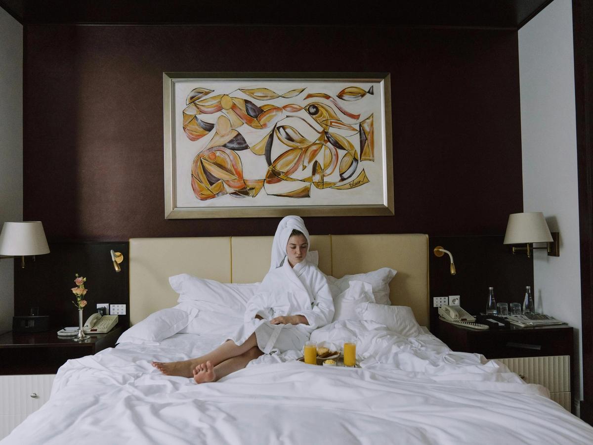 A woman relaxes in a plush hotel bed wearing a robe and towel wrap, with breakfast served on a tray beside her. The image reflects the calm, personalized comfort that boutique hotels are known for.