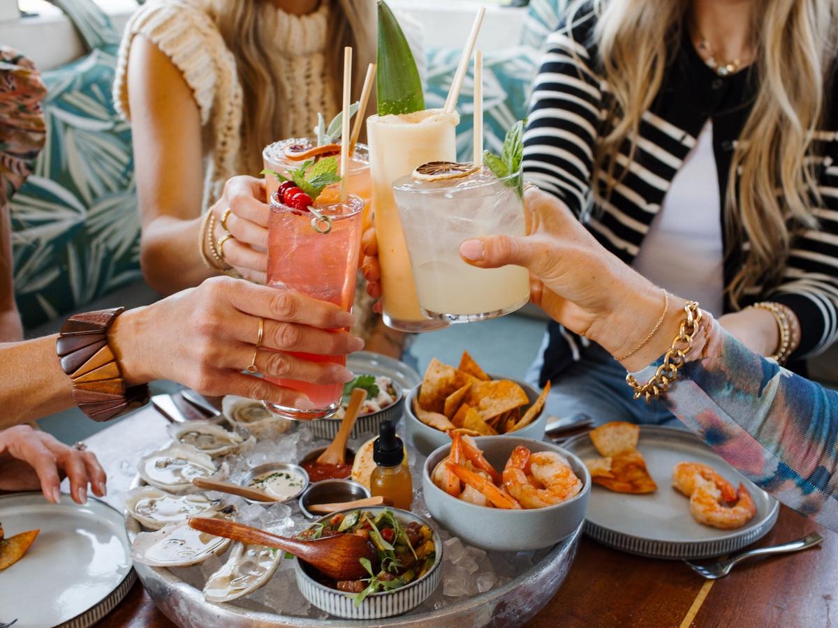 Friends clink cocktails over a shared seafood platter at The Longboard, where coastal bites meet tropical drinks. It’s the kind of laid-back place where every meal feels like a celebration.