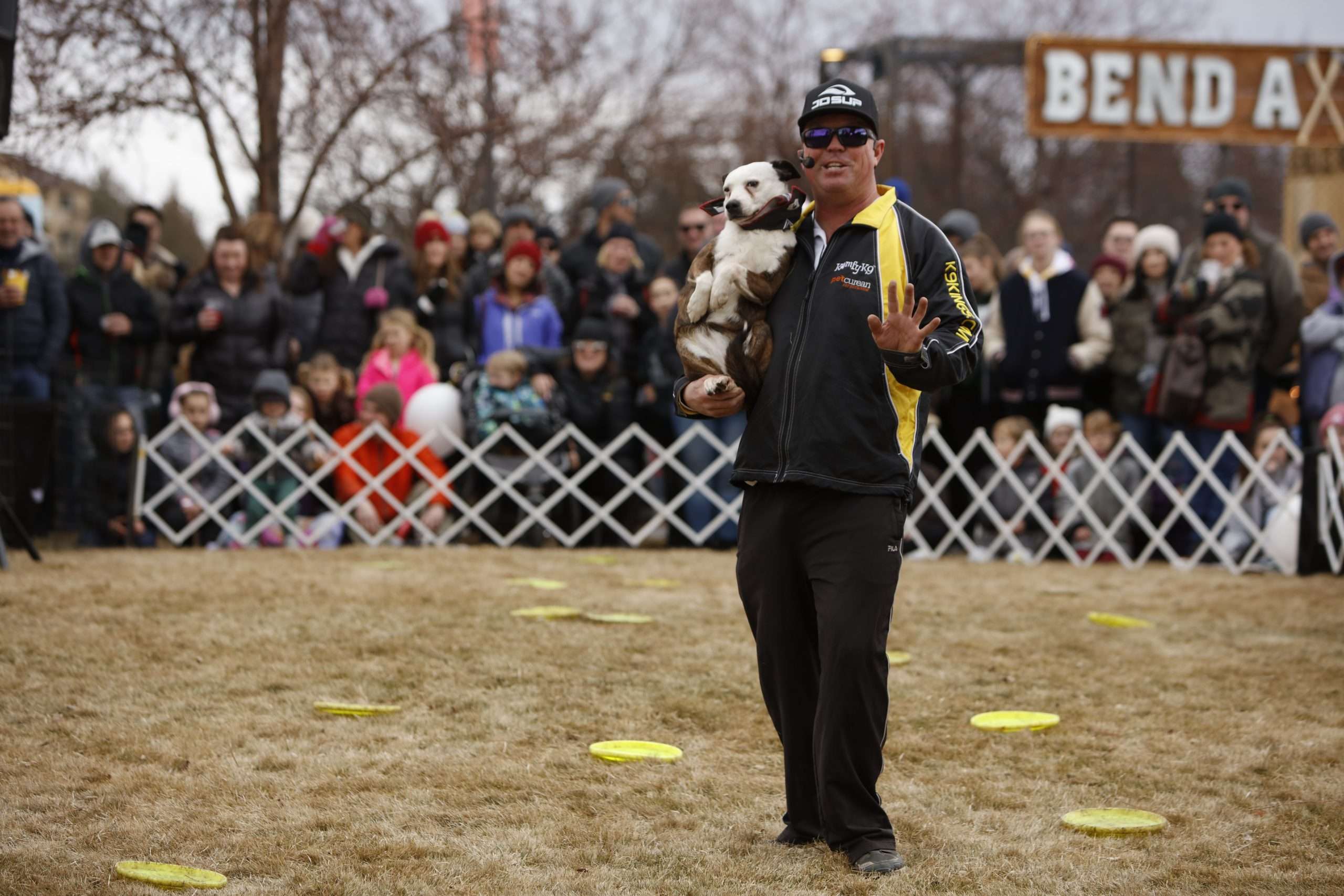 A lively crowd watches a man and his dog perform at an outdoor winter show surrounded by frisbees on the ground. Families bundled up in winter coats enjoy the entertainment at Bend’s WinterFest celebration.