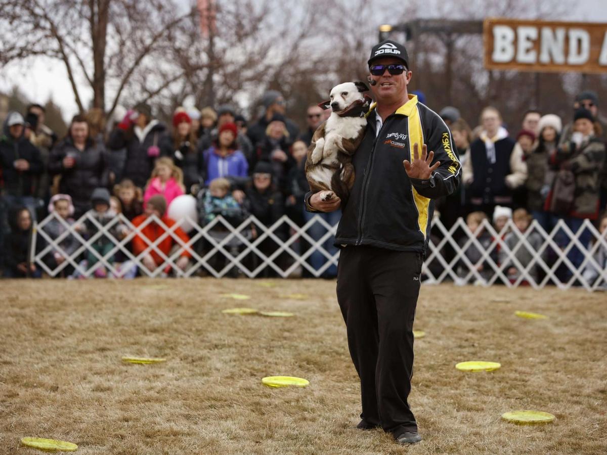 A lively crowd watches a man and his dog perform at an outdoor winter show surrounded by frisbees on the ground. Families bundled up in winter coats enjoy the entertainment at Bend’s WinterFest celebration.