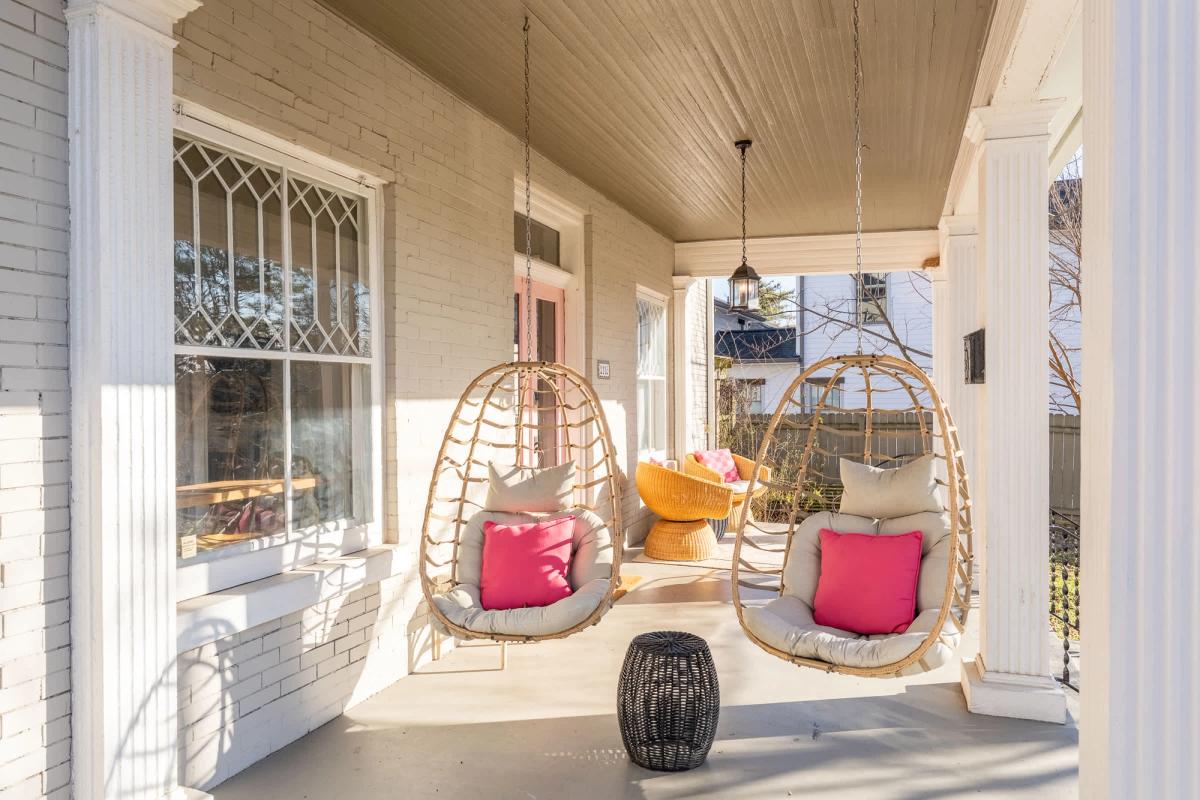 A charming Southern front porch with hanging wicker egg chairs, pink accent pillows, white columns, and soft natural light, creating an inviting spot to relax at a Nashville retreat.