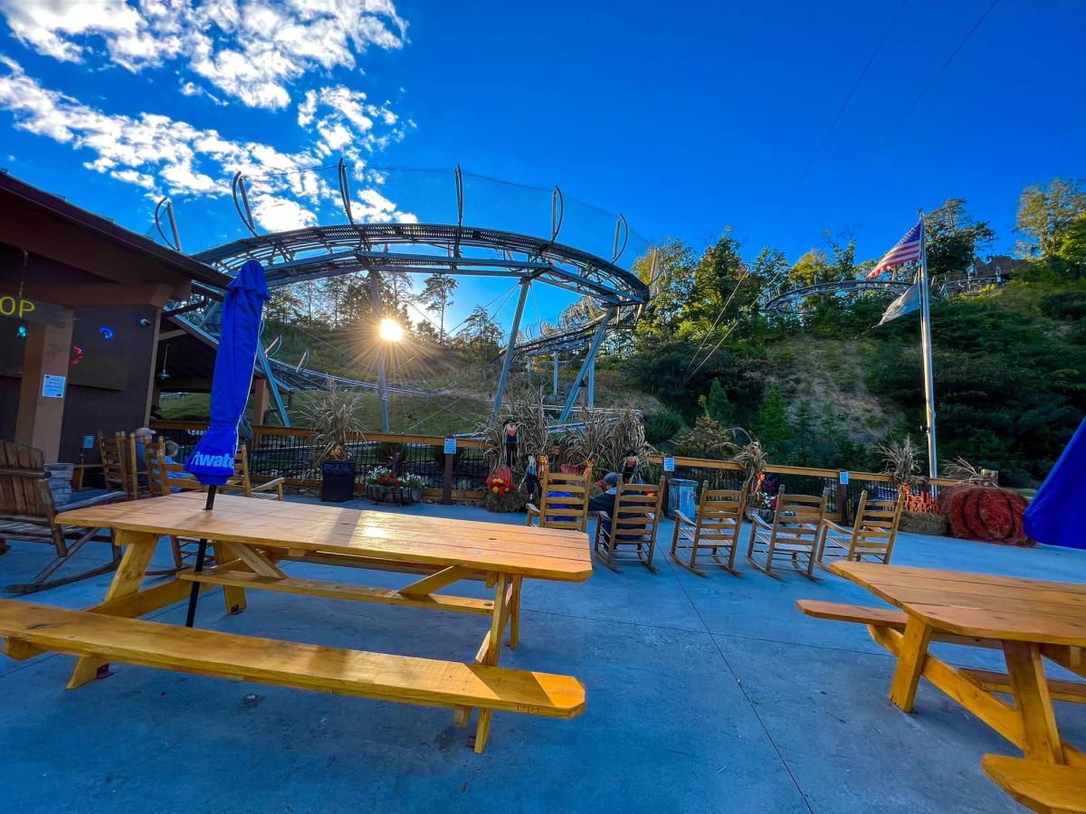 A sunny day at a Smoky Mountain coaster park featuring wooden picnic tables and rocking chairs under a clear blue sky. The metal coaster track winds above the seating area as the sun shines through the trees.