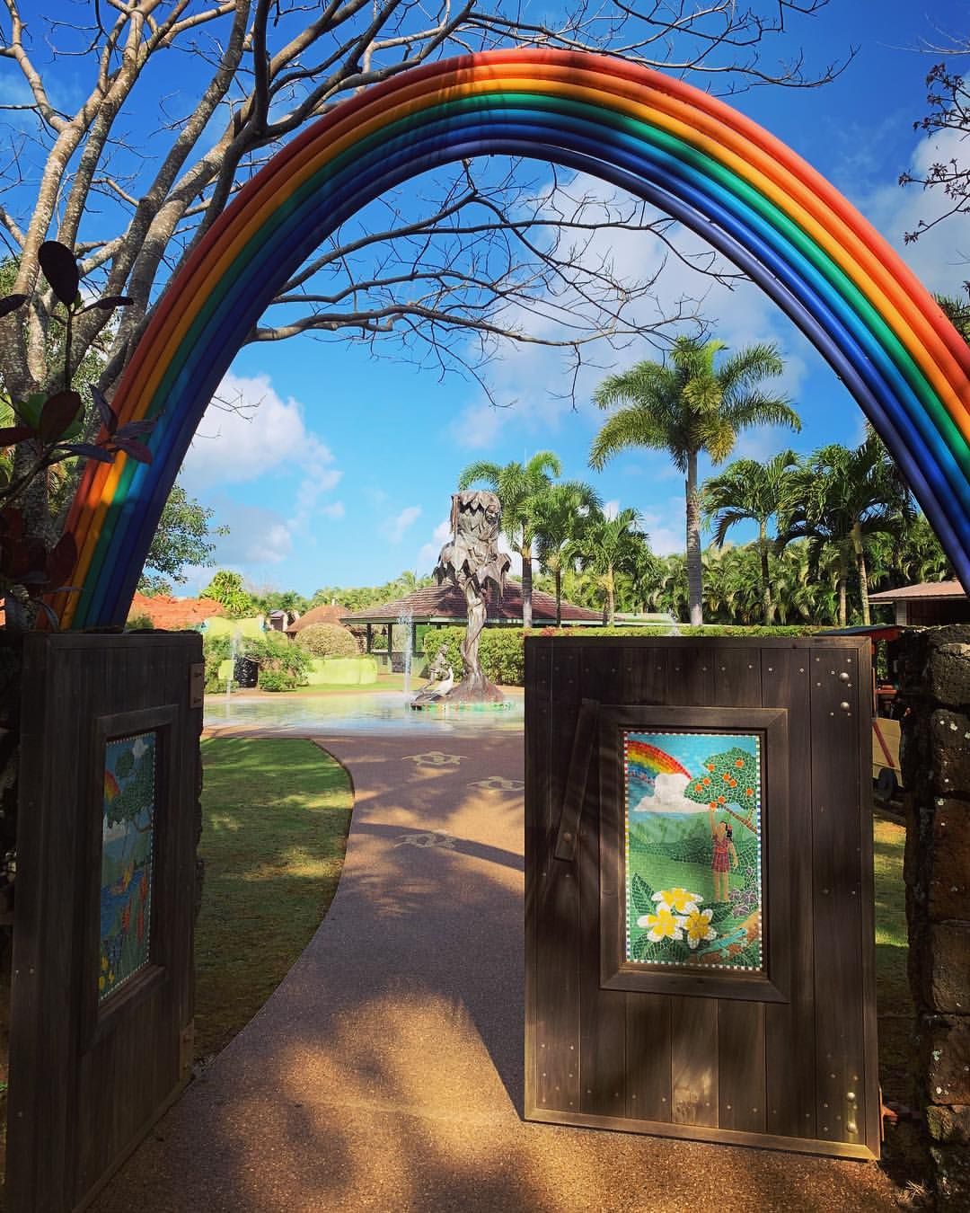 A vibrant rainbow arch gateway welcomes visitors into Na ʻAina Kai Botanical Gardens and Sculpture Park in Kauai, framing a path leading to a dramatic tree sculpture and fountain surrounded by tropical palms.