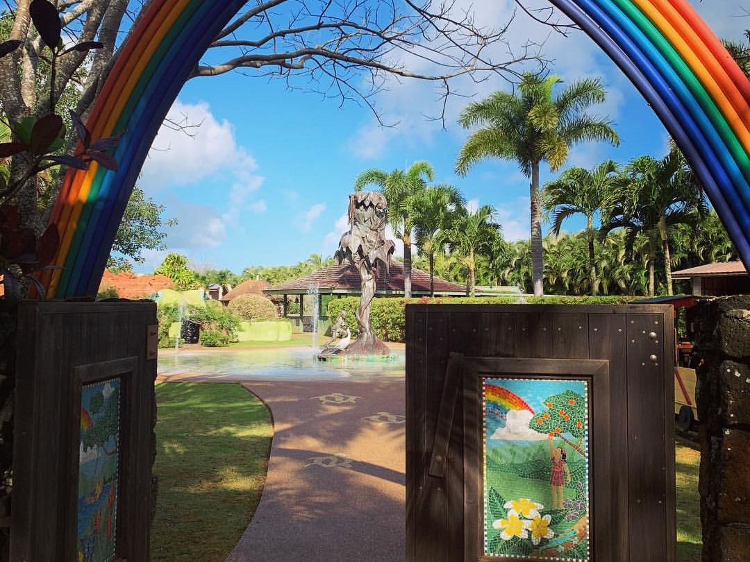 A vibrant rainbow arch gateway welcomes visitors into Na ʻAina Kai Botanical Gardens and Sculpture Park in Kauai, framing a path leading to a dramatic tree sculpture and fountain surrounded by tropical palms.