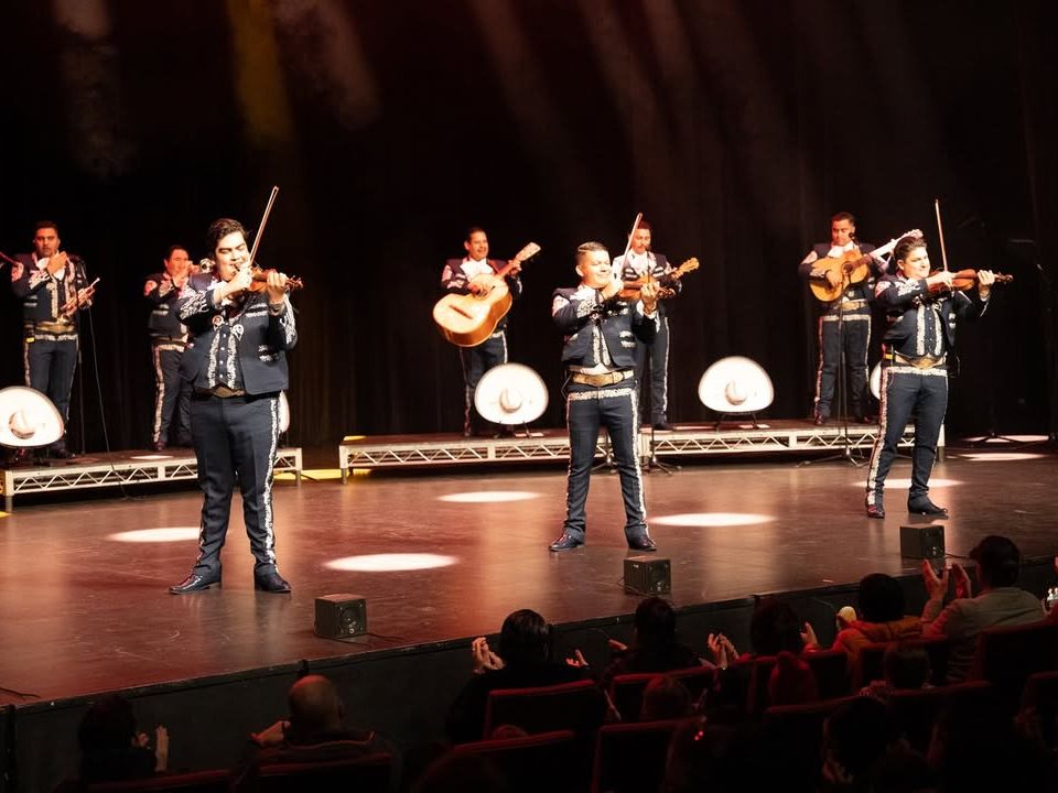 A mariachi band performs live on stage at the Center for the Arts in Jackson Hole, wearing traditional outfits and playing violins and guitars. The audience watches closely as the venue hosts a cultural music performance known for its strong acoustics and intimate seating.