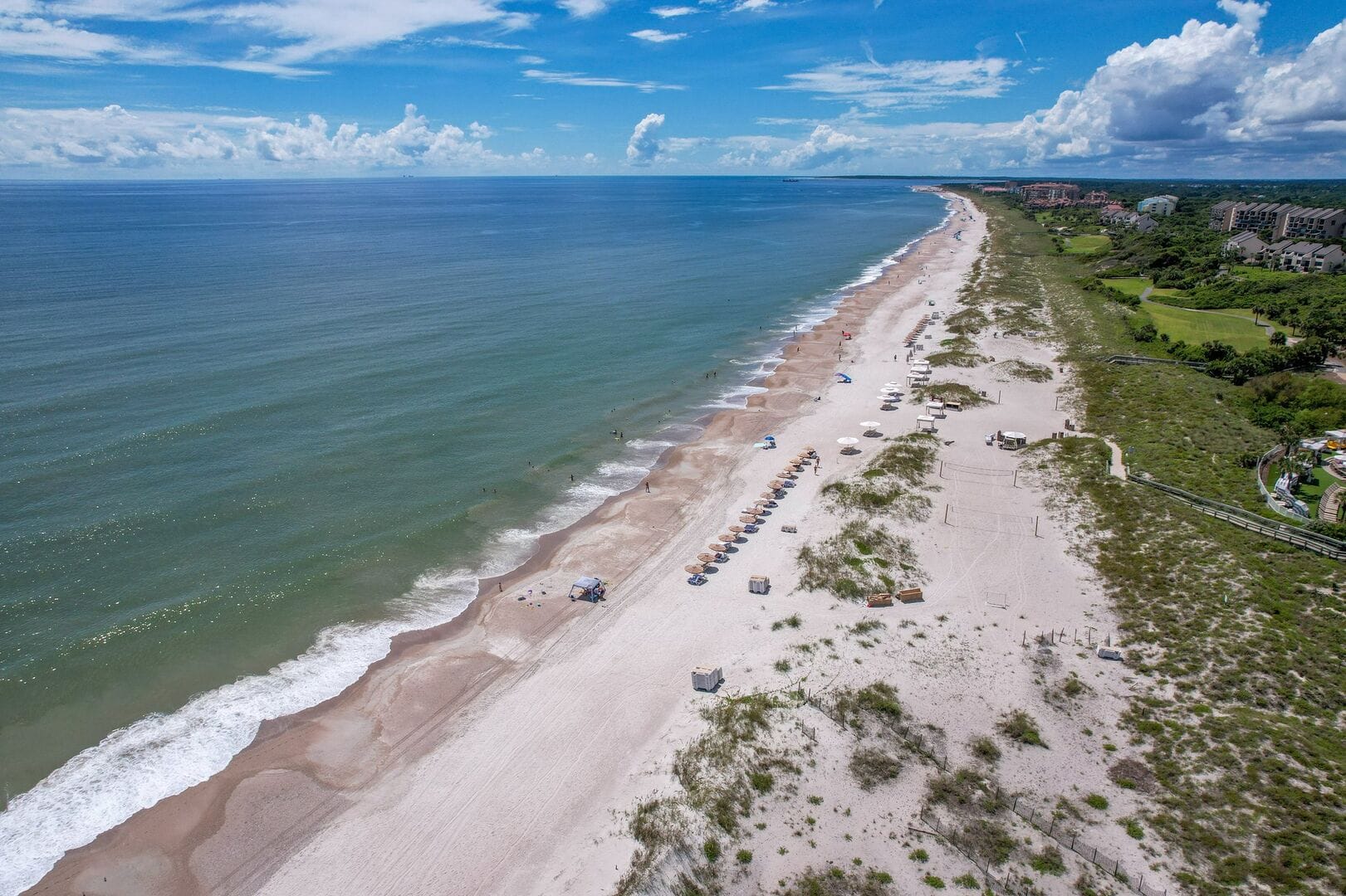An aerial view of Amelia Island, Florida, shows wide sandy beaches meeting calm blue waters along the Atlantic coast. Beach umbrellas line the shore near green dunes and resort properties. Amelia Island is known for its peaceful beaches and natural coastal beauty.