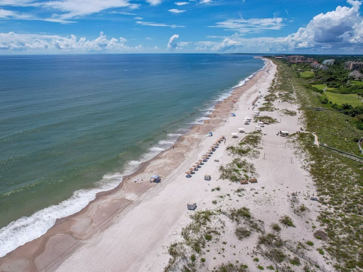 An aerial view of Amelia Island, Florida, shows wide sandy beaches meeting calm blue waters along the Atlantic coast. Beach umbrellas line the shore near green dunes and resort properties. Amelia Island is known for its peaceful beaches and natural coastal beauty.