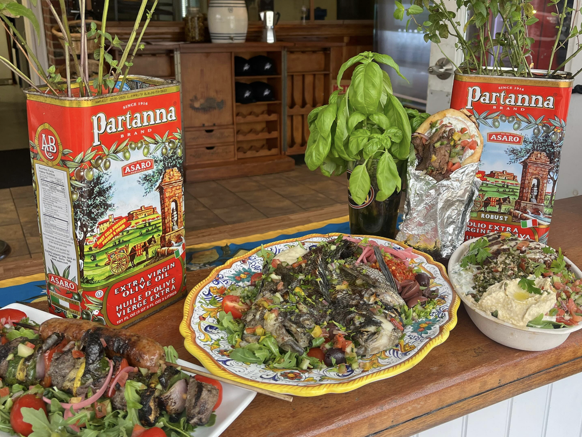 Mediterranean feast display featuring grilled fish on a decorative platter, skewers with vegetables, a gyro wrapped in foil, and a bowl of hummus and tabbouleh, surrounded by fresh herbs and vintage Partanna olive oil tins.