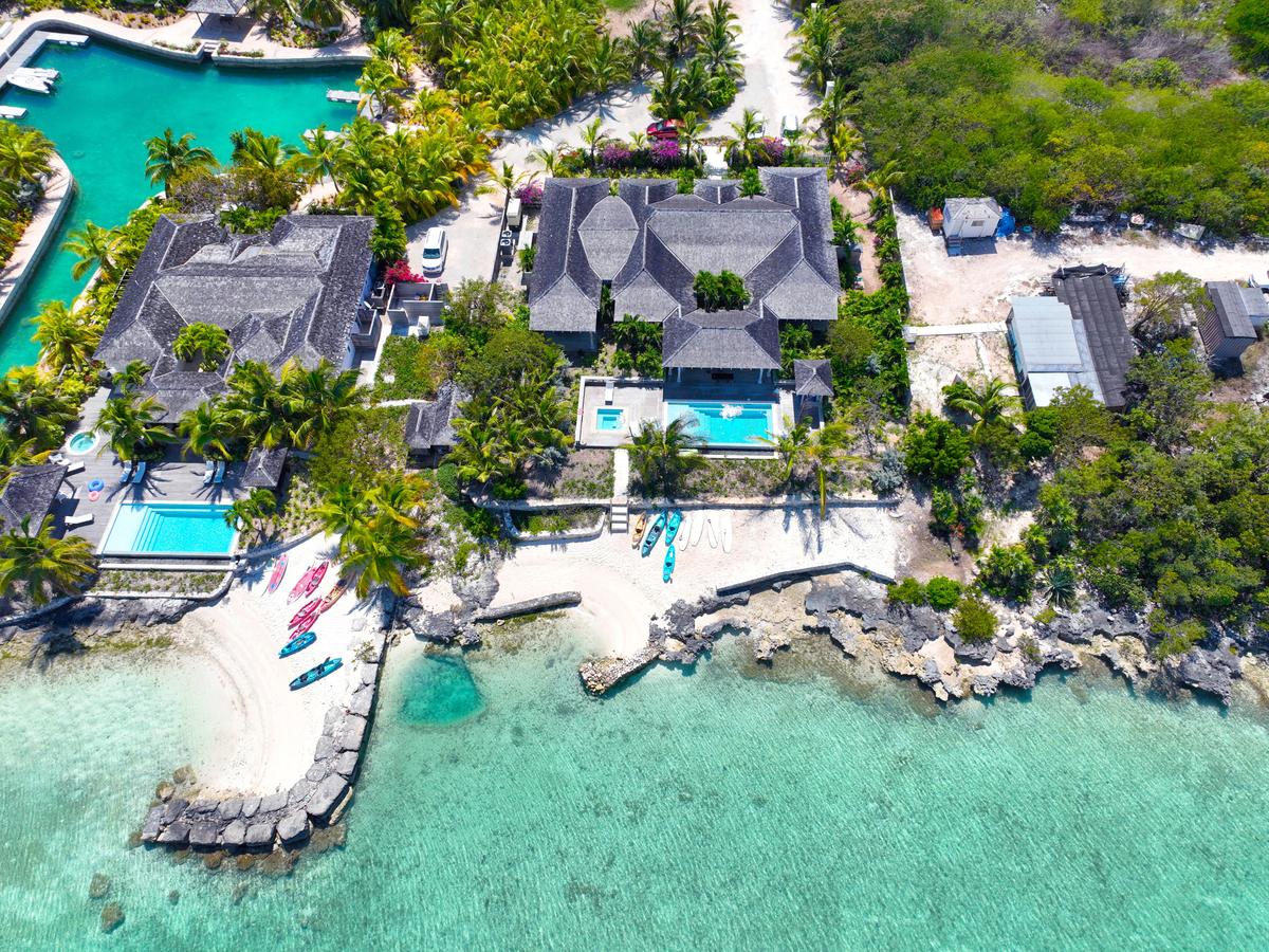 Aerial view of a luxury beachfront villa with thatched roofs, private pools, and kayaks lined up along a white sandy shore beside clear turquoise water.