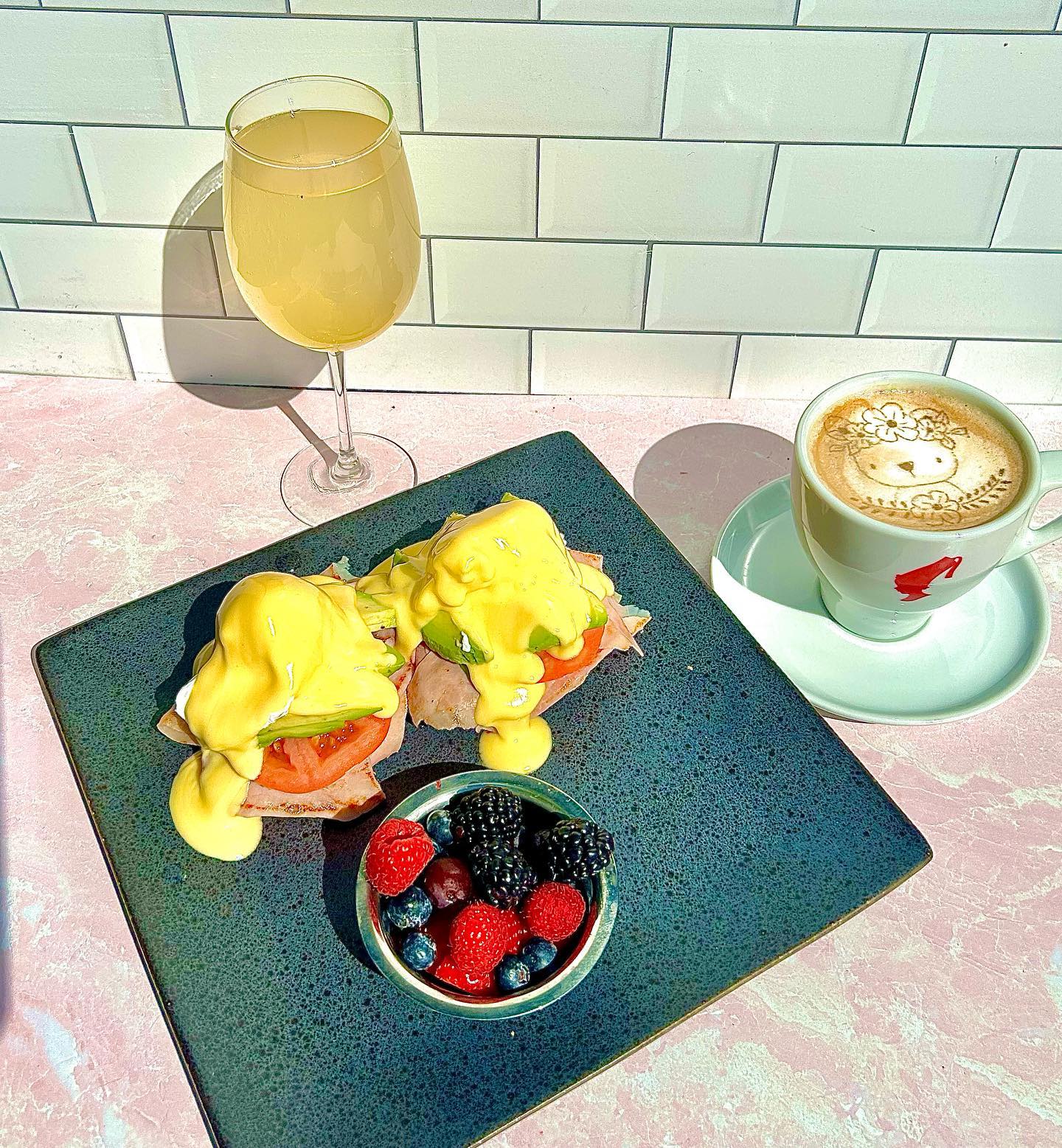 A bright brunch plate with eggs benedict, fresh berries, and a latte sits under soft sunlight, giving the whole scene a warm, cheerful vibe. The creamy hollandaise and fresh fruit make the meal look rich and satisfying. It’s the kind of breakfast that feels slow, cozy, and perfect for a relaxed morning.