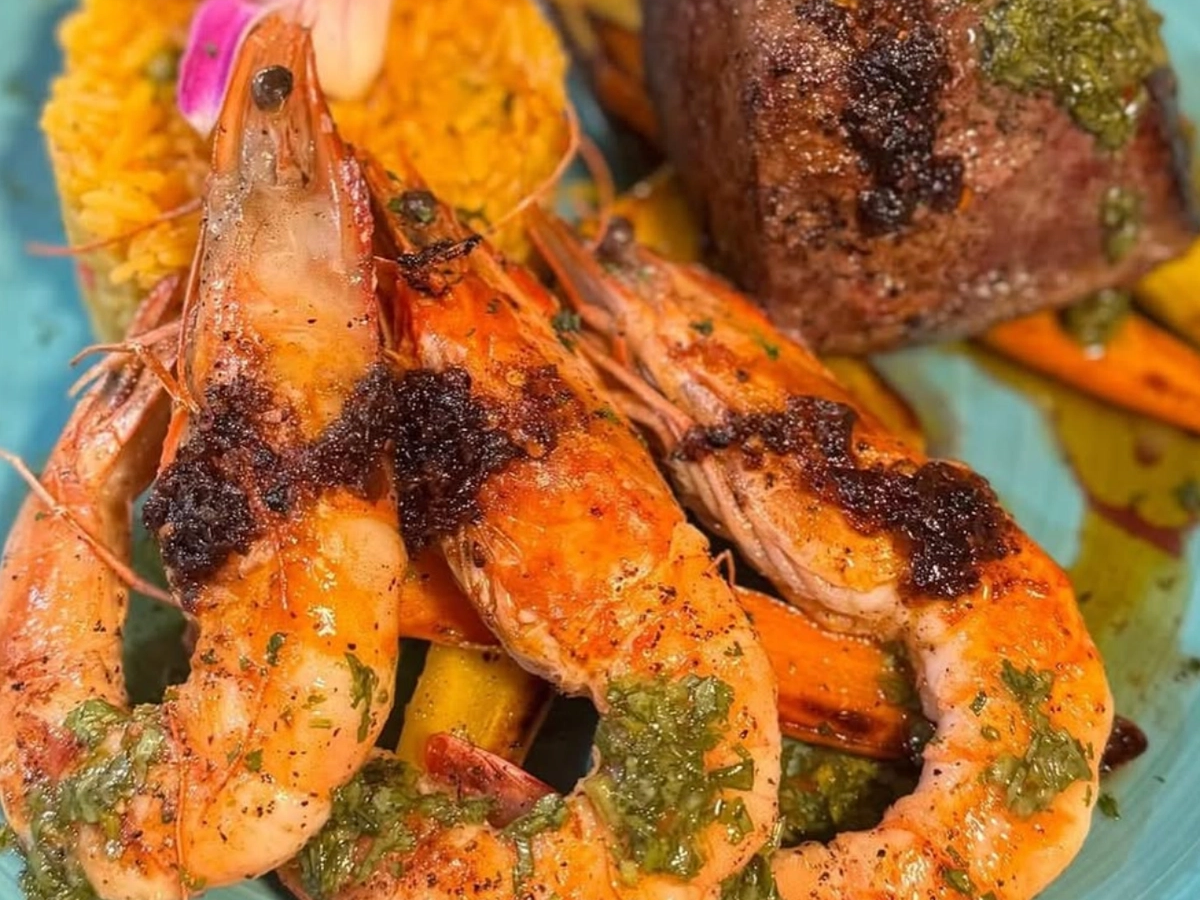 Vibrant surf-and-turf dish featuring grilled shrimp with chimichurri, a seared filet mignon, yellow rice, and roasted vegetables, garnished with a tropical flower.