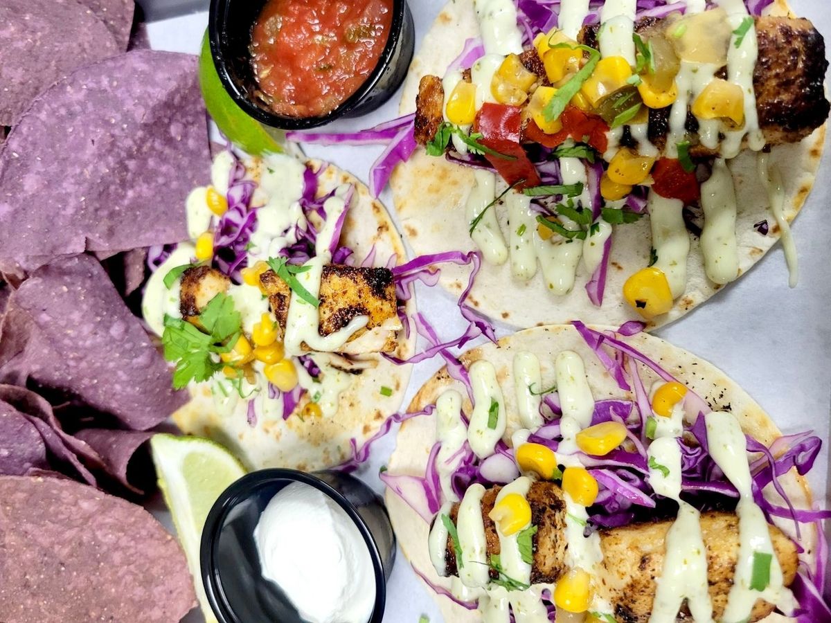 Three fish tacos topped with corn salsa, purple cabbage, and creamy sauce, served with salsa, sour cream, and purple chips. A fresh and bold beachside meal.