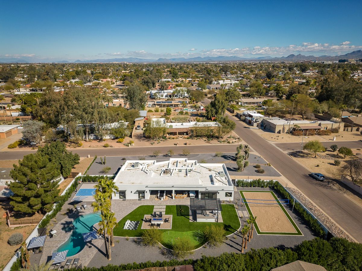 An expansive aerial view of a luxury desert estate featuring a resort-style backyard with a pool, outdoor dining pavilion, sand volleyball court, and manicured lawn, all set against sweeping mountain vistas.