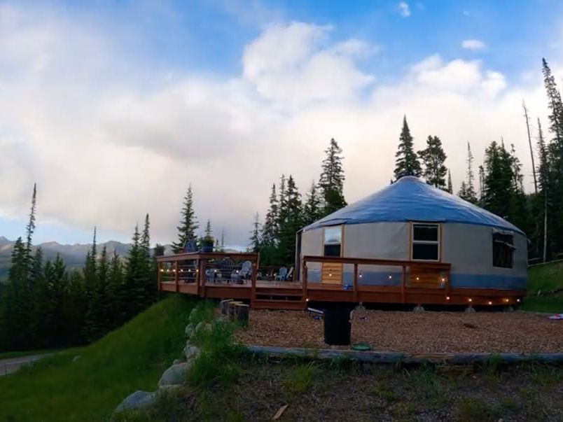 A cozy mountain yurt with a wraparound deck sits among tall pines in Big Sky, Montana. Guests can enjoy unique dining experiences here with panoramic views of the mountains and a warm, rustic vibe.