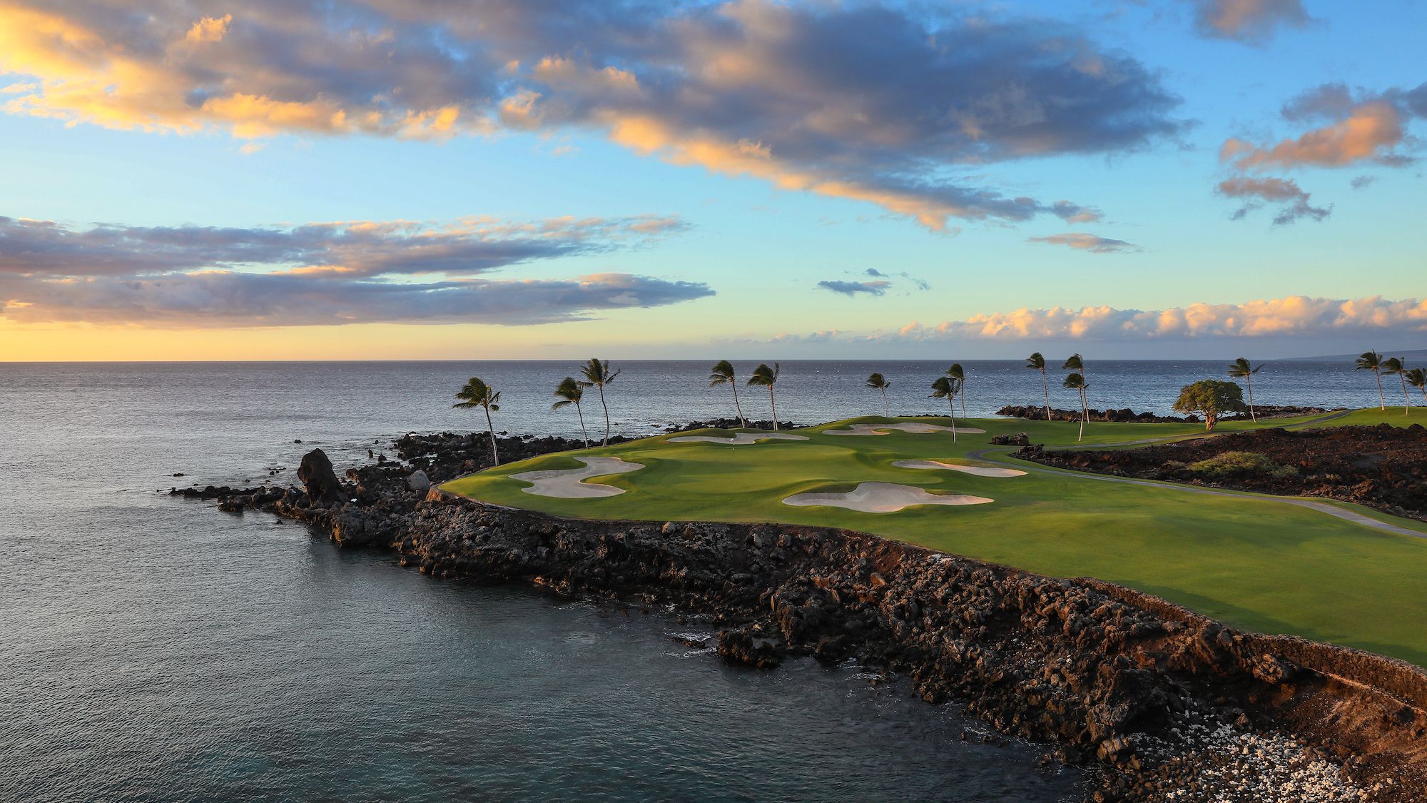The dramatic oceanfront holes of the Mauna Lani South Course on the Big Island of Hawaii jut out over black lava rock into the Pacific, with swaying palm trees and a fiery sunset sky reflected across the calm blue sea. 