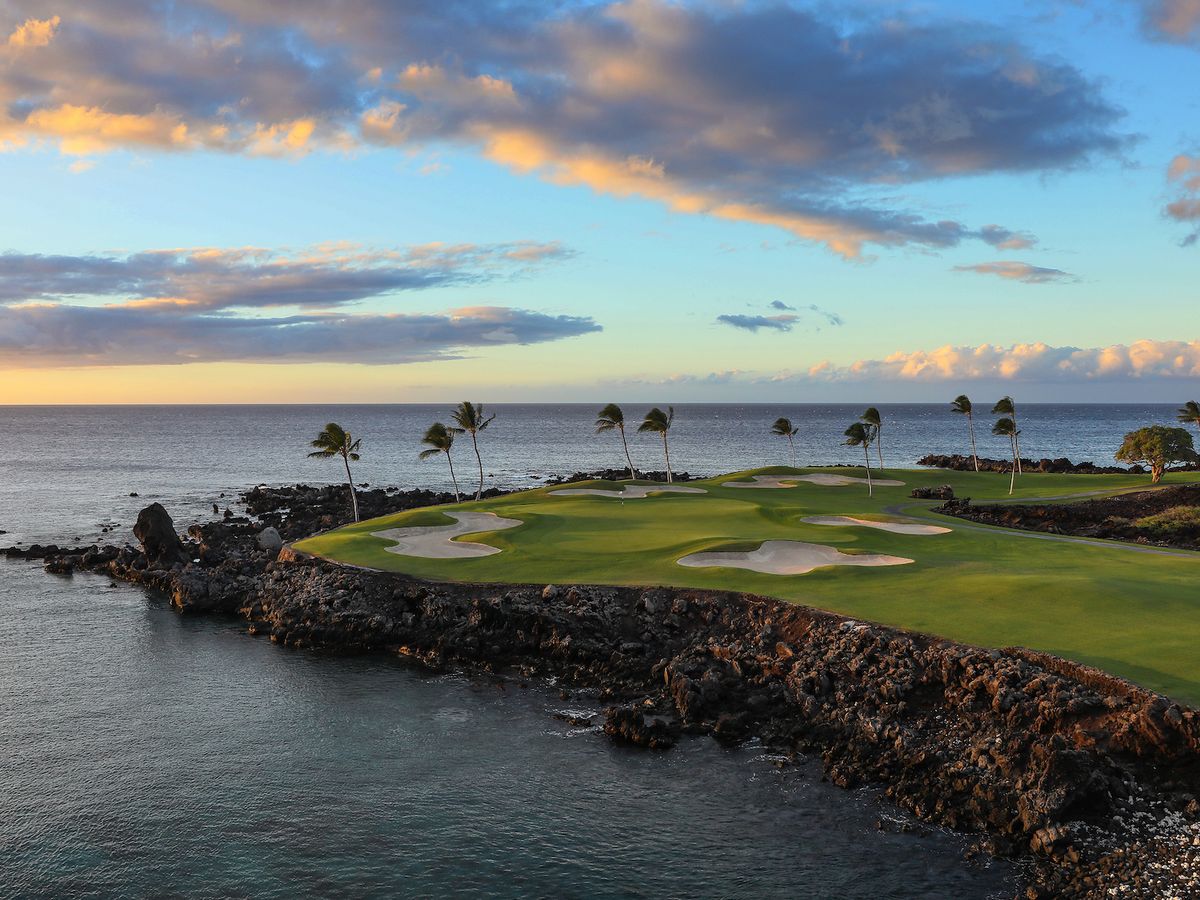 The dramatic oceanfront holes of the Mauna Lani South Course on the Big Island of Hawaii jut out over black lava rock into the Pacific, with swaying palm trees and a fiery sunset sky reflected across the calm blue sea.