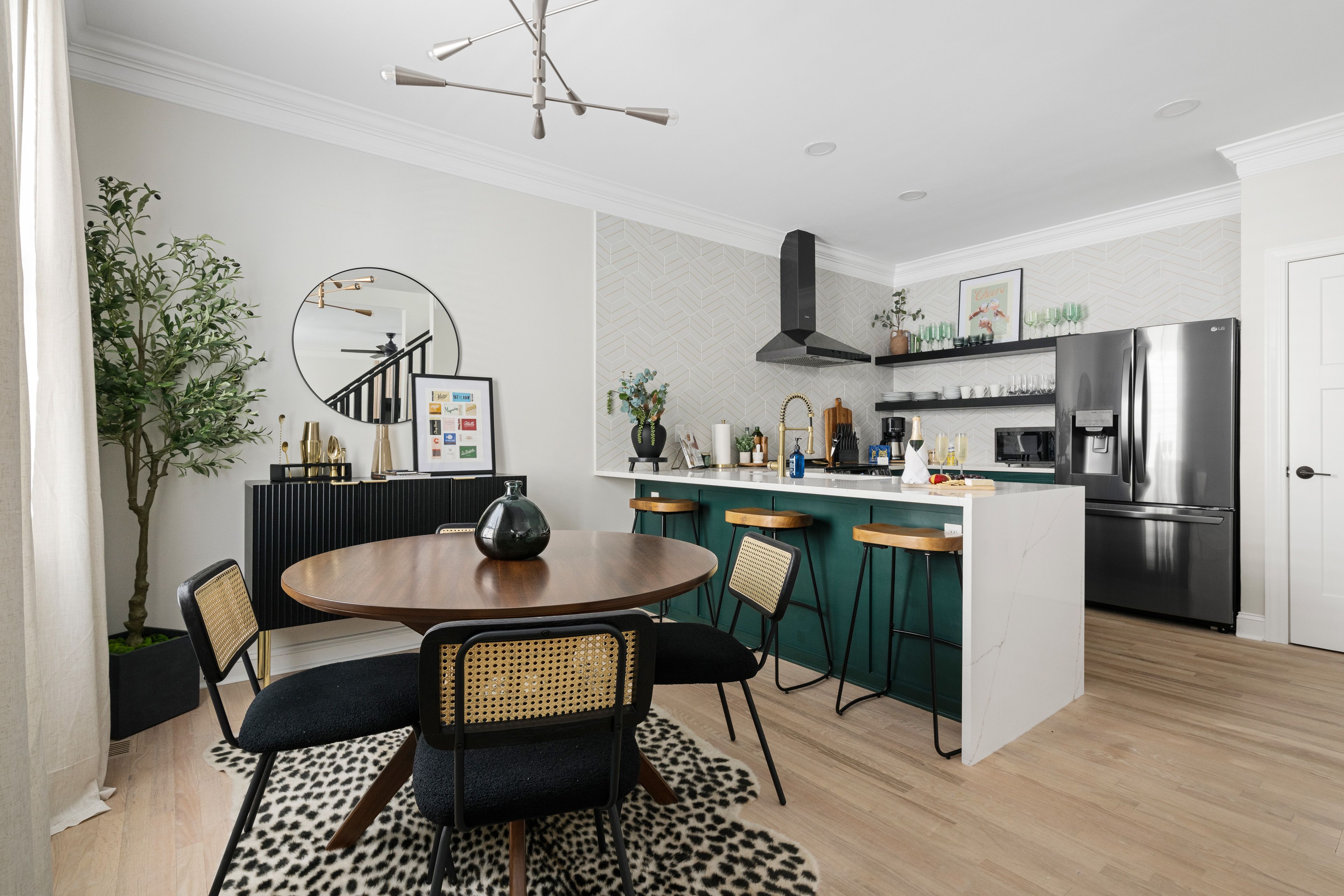 A modern kitchen and dining area features a bold green island with wood barstools, a round dining table with rattan-back chairs, open shelving, and stylish accents like a black fridge and geometric wallpaper.