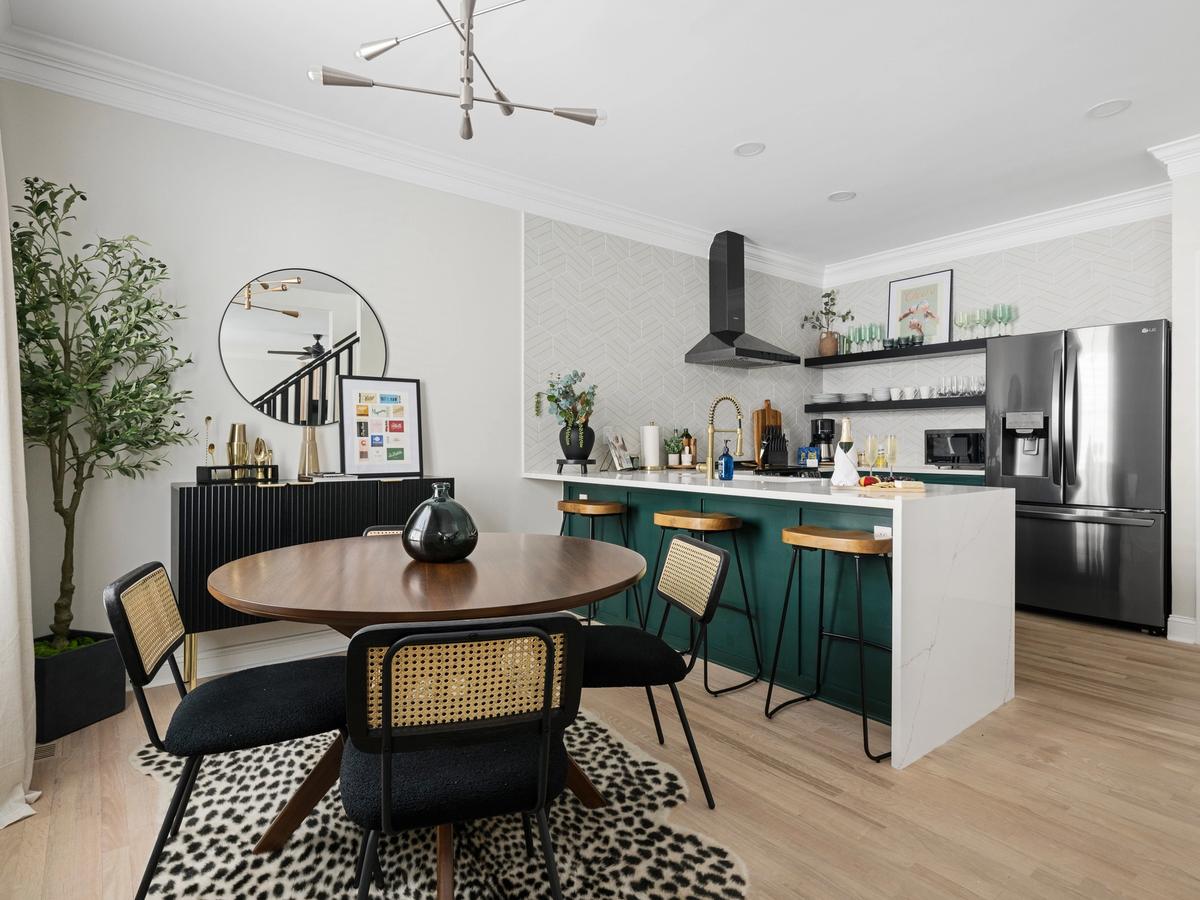 A modern kitchen and dining area features a bold green island with wood barstools, a round dining table with rattan-back chairs, open shelving, and stylish accents like a black fridge and geometric wallpaper.