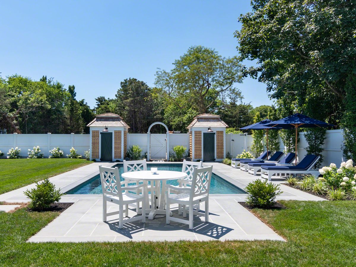 A stylish backyard pool area features white patio furniture, navy blue lounge chairs with matching umbrellas, manicured landscaping, and two charming brick pool houses against a white privacy fence.