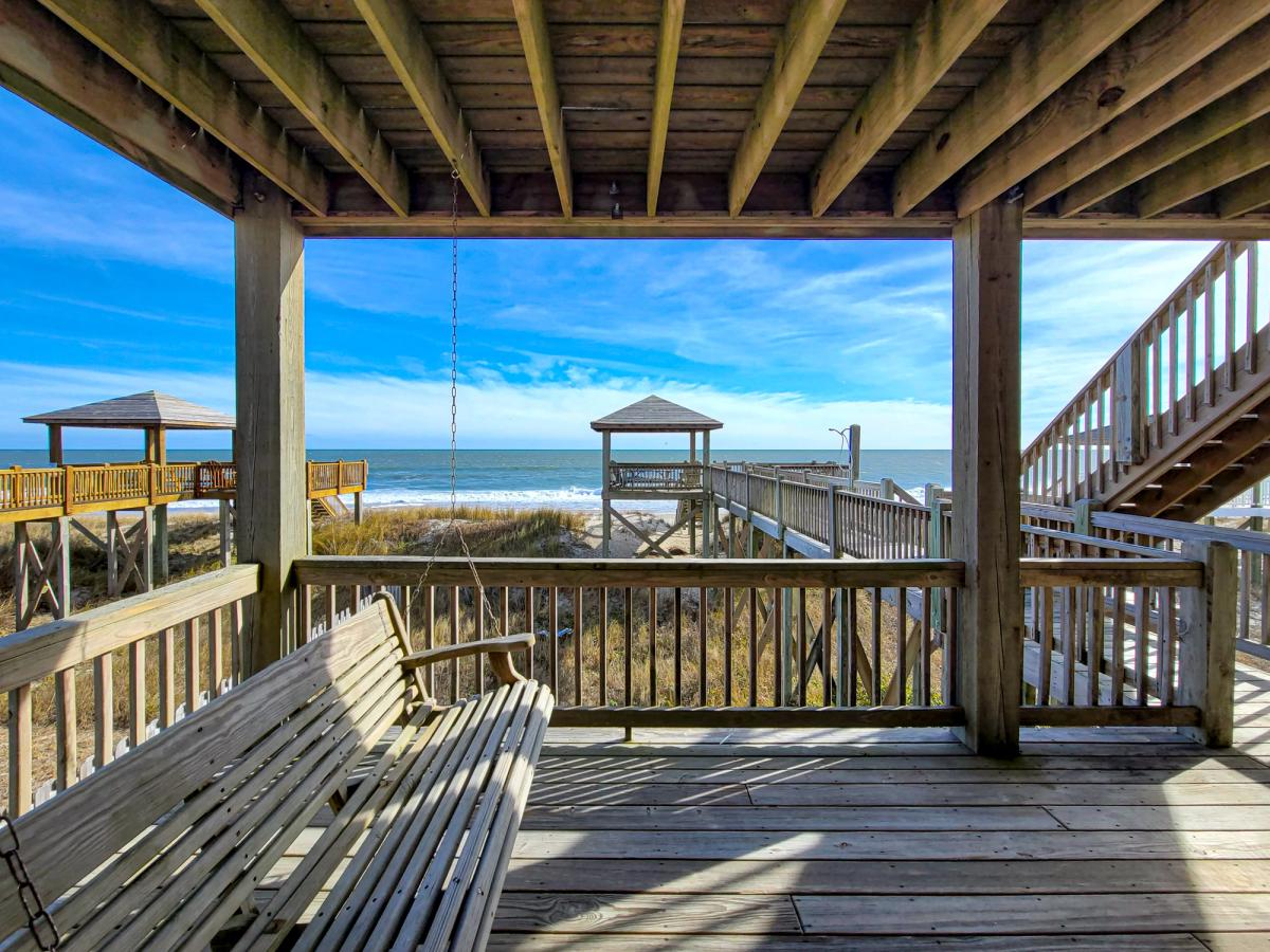 A row of coastal blue townhomes with wooden staircases leading up to elevated decks, surrounded by palm trees, white gravel landscaping, and driveways with recycling bins.         You said:   ChatGPT said: Alt text: A shaded beachfront porch with a wooden porch swing, overlooking a private boardwalk leading to a gazebo and the ocean beyond under a clear blue sky.