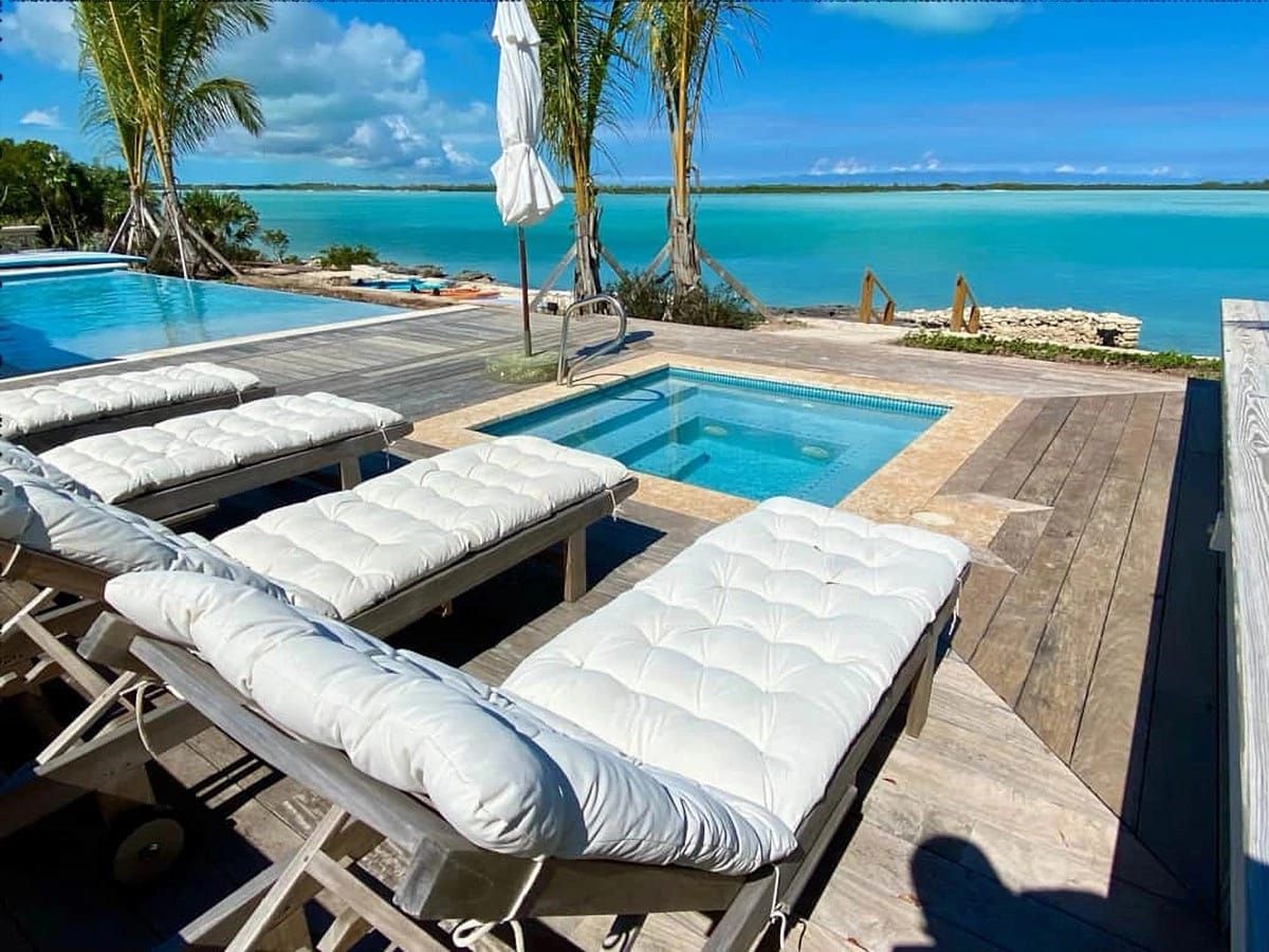 Plush white lounge chairs line a wooden deck beside a hot tub and infinity pool, overlooking turquoise ocean waters under a bright blue sky.