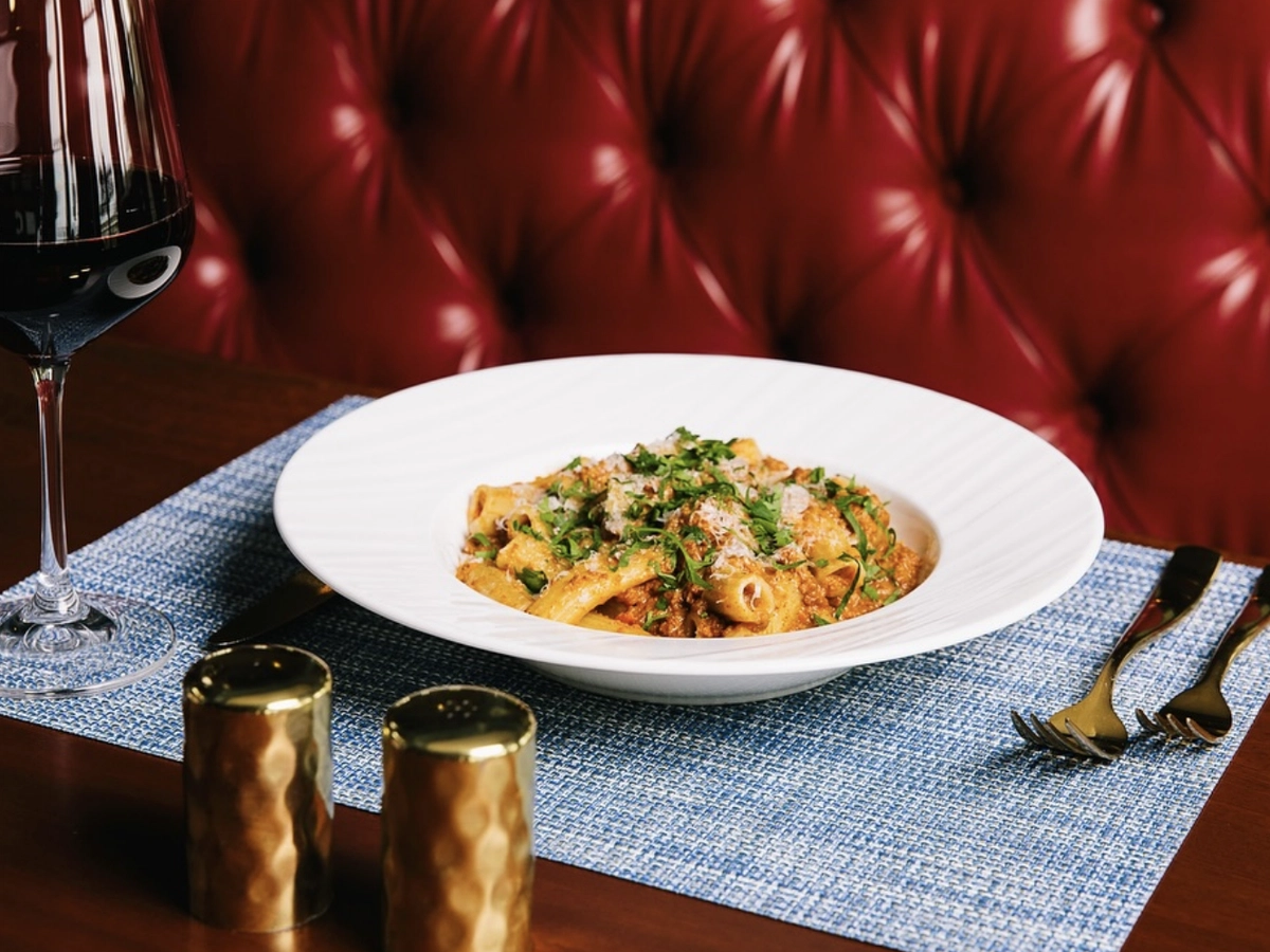 Elegant bowl of rigatoni pasta in a rich meat sauce, garnished with fresh herbs and Parmesan, served with a glass of red wine on a blue placemat in a cozy red leather booth setting.