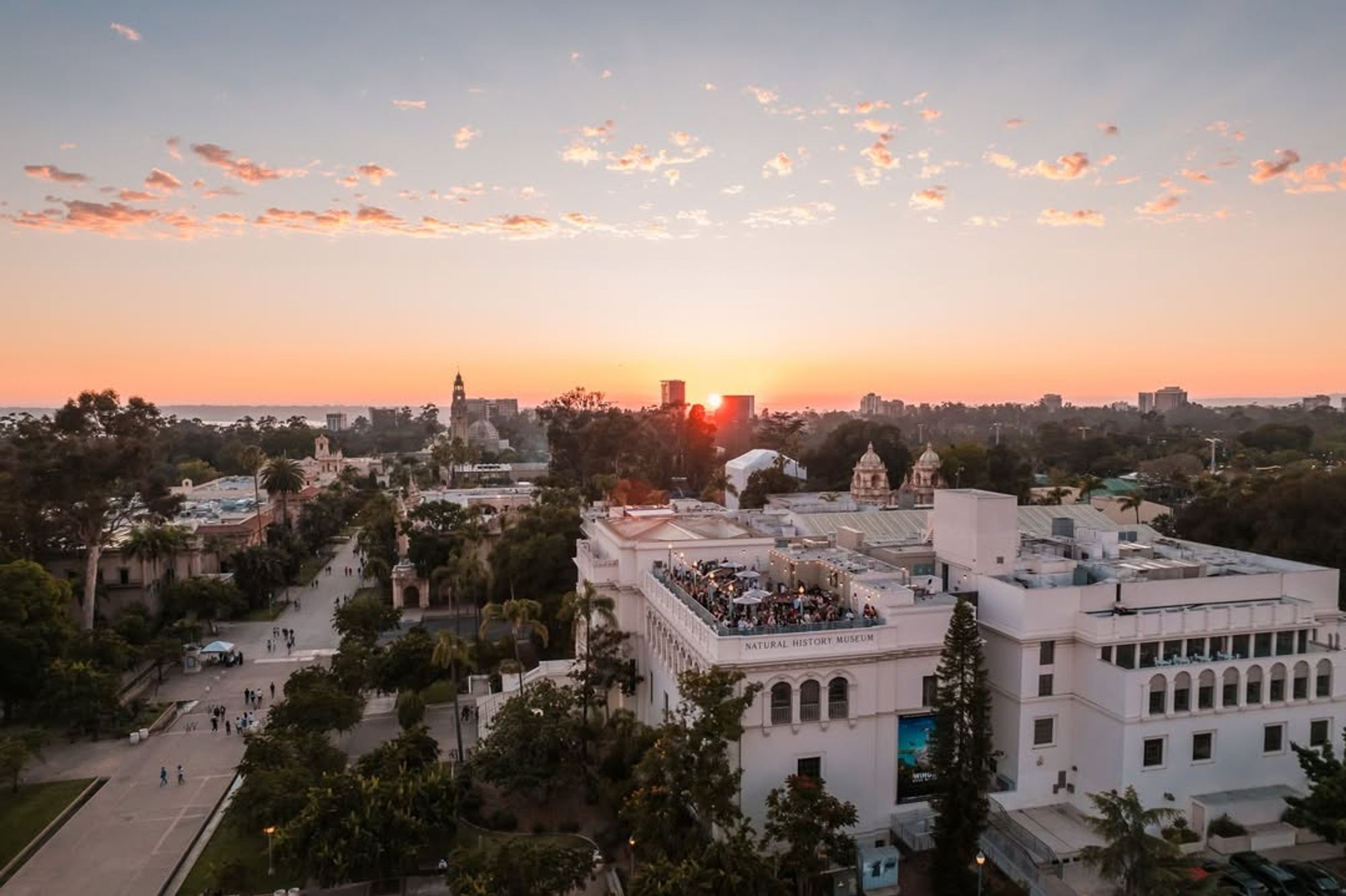 Sunset glows over Balboa Park’s museums and historic buildings, with crowds enjoying a rooftop event at the Natural History Museum. The view captures the charm of this iconic San Diego destination.