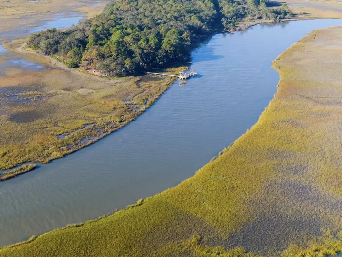 Aerial view of a winding tidal creek bordered by golden marsh grasses and leading to a forested island with a dock and boat.