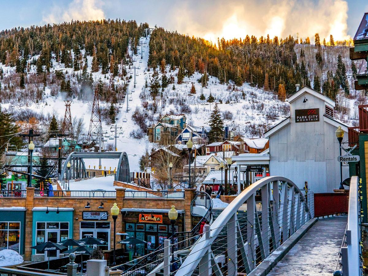 A snowy mountain scene in Park City, Utah, with ski lifts ascending the slope and a charming town below filled with colorful buildings, shops, and bridges.