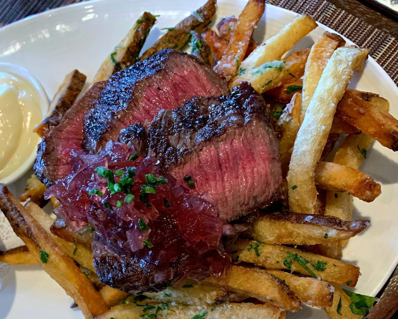 Slices of medium-rare steak served on top of crispy herb-seasoned fries, topped with caramelized onions and a side of aioli.