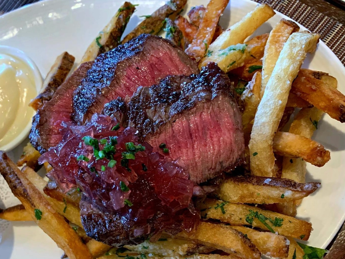Slices of medium-rare steak served on top of crispy herb-seasoned fries, topped with caramelized onions and a side of aioli.
