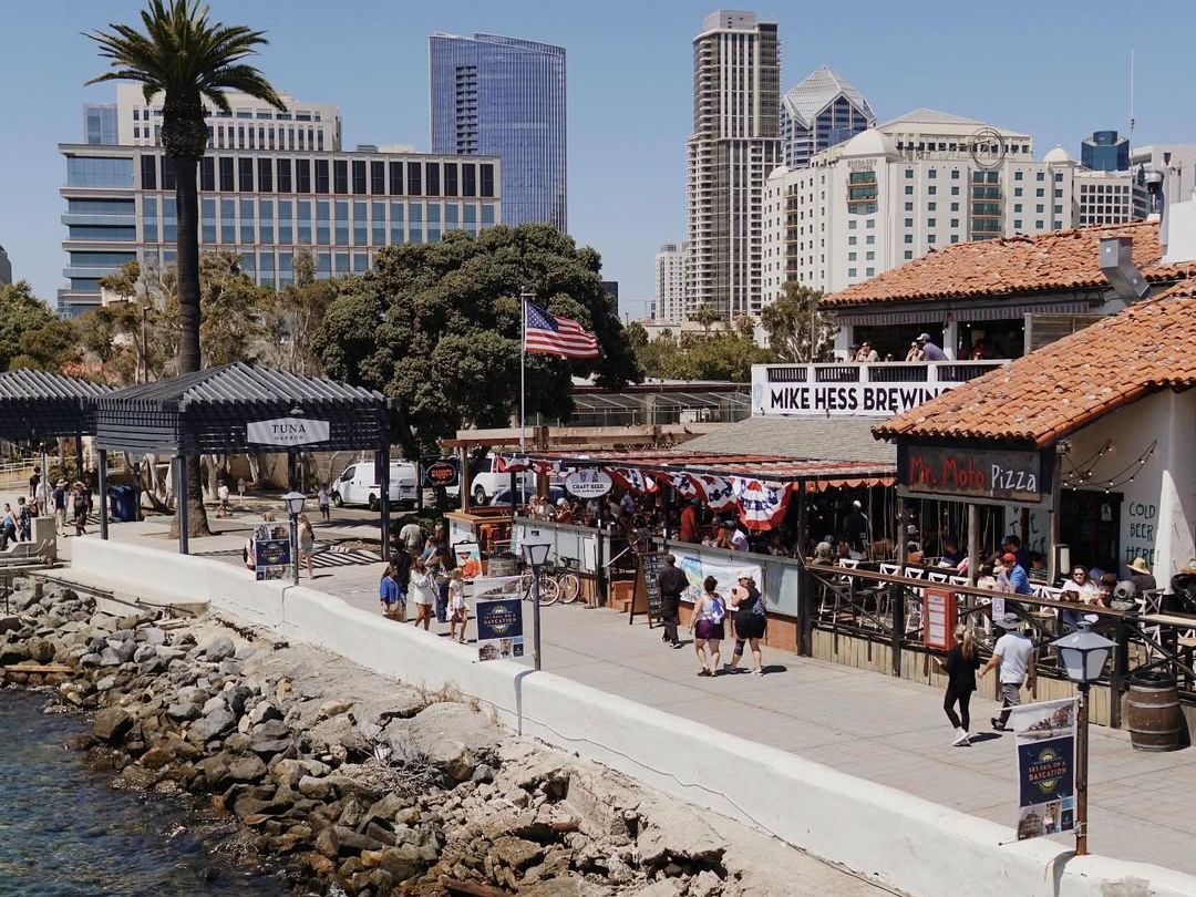 A lively waterfront scene at Seaport Village in San Diego, with people strolling along the boardwalk, colorful shops, and modern high-rise buildings in the background. The area blends coastal charm with city views, making it a favorite spot for dining and sightseeing.