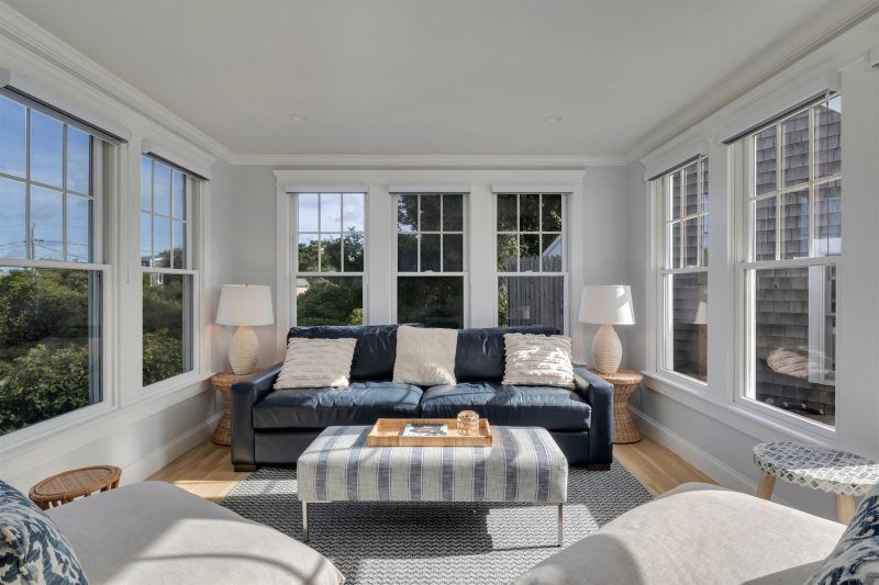 Sunlit sitting room with wraparound windows, a navy leather sofa accented with neutral pillows, woven side tables with ceramic lamps, and a striped ottoman creating a bright, coastal-inspired retreat.
