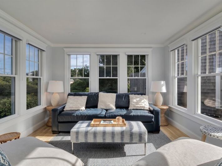 Sunlit sitting room with wraparound windows, a navy leather sofa accented with neutral pillows, woven side tables with ceramic lamps, and a striped ottoman creating a bright, coastal-inspired retreat.
