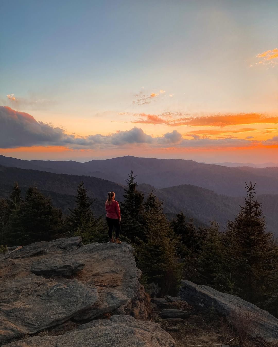 A hiker stands on a rocky overlook at Mount Mitchell, watching the sun set over layers of blue mountain ridges. This peaceful sunset view shows why Mount Mitchell is a favorite spot for scenic hikes and breathtaking mountain views in North Carolina.