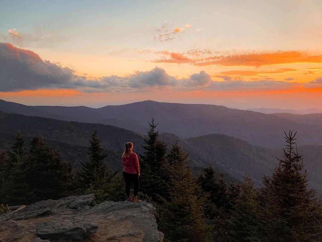 A hiker stands on a rocky overlook at Mount Mitchell, watching the sun set over layers of blue mountain ridges. This peaceful sunset view shows why Mount Mitchell is a favorite spot for scenic hikes and breathtaking mountain views in North Carolina.