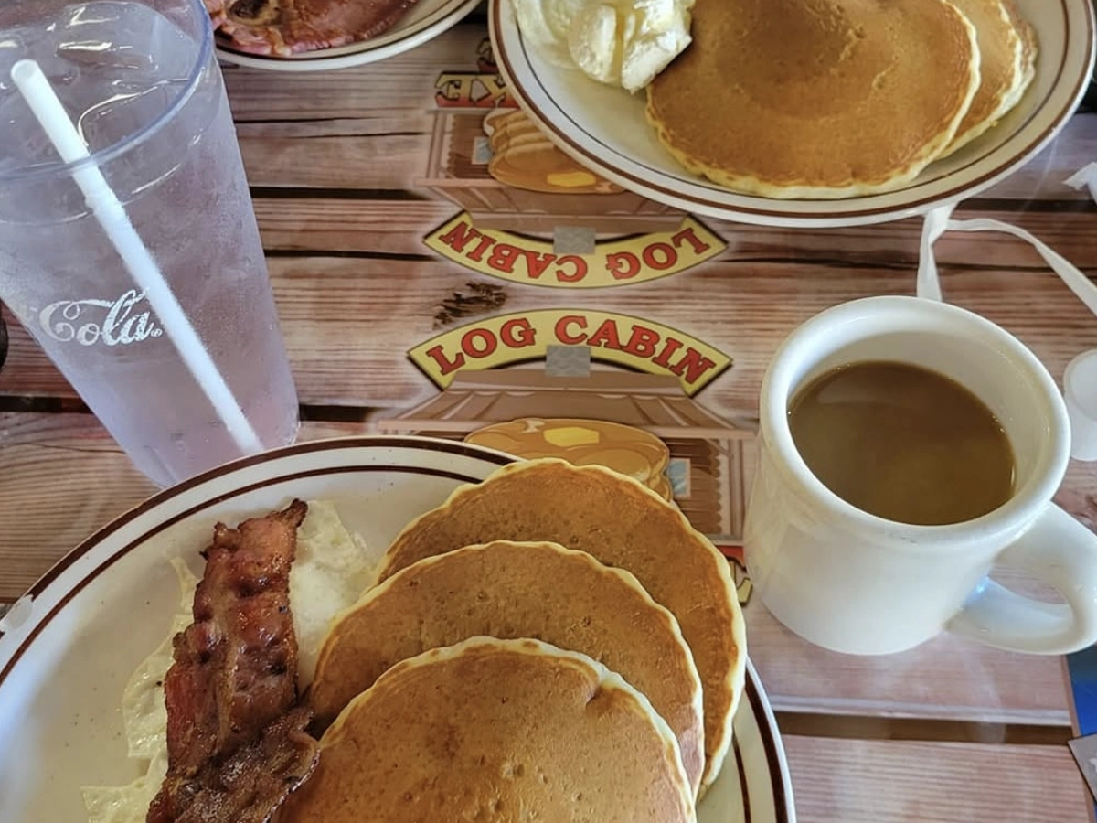 A hearty breakfast spread featuring two plates of pancakes with eggs and bacon or ham, a mug of coffee, and a glass of ice water on a table with a "Log Cabin" diner-themed placemat.