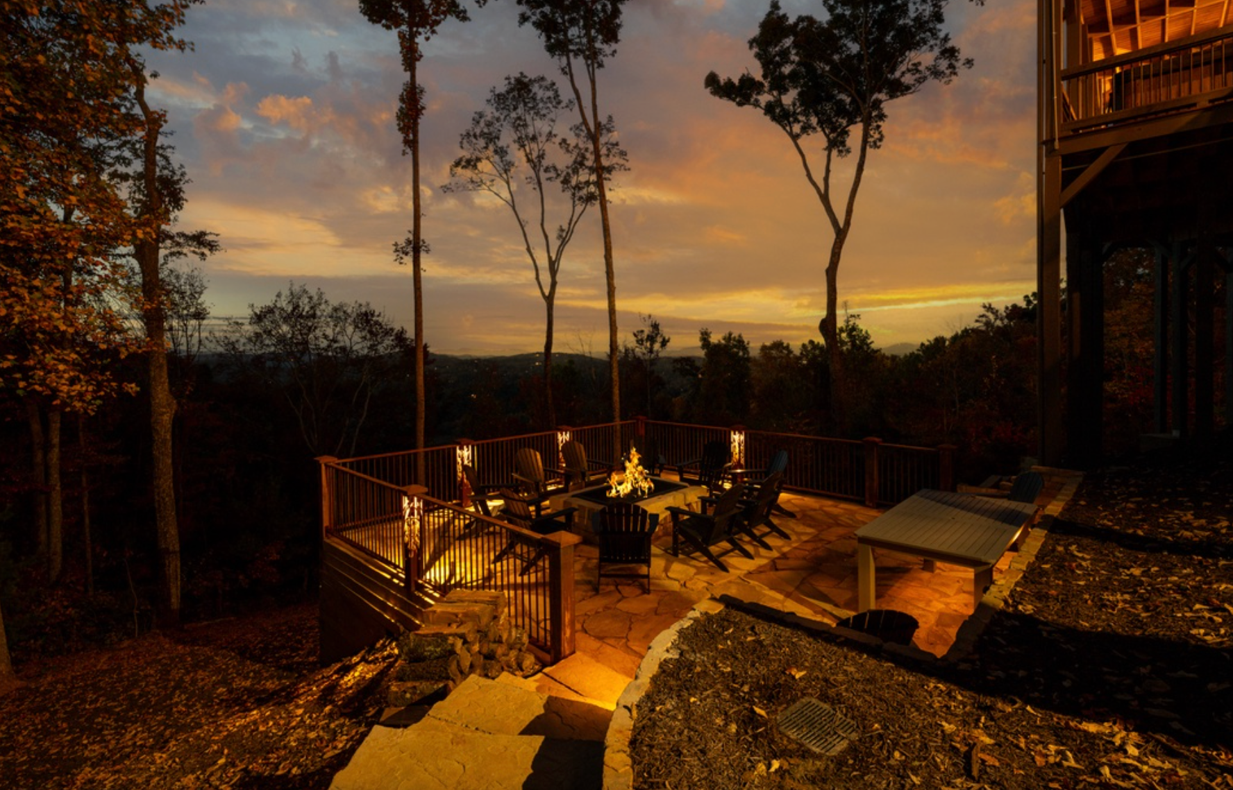 Outdoor patio with a fire pit and seating area overlooking a wooded mountain landscape at sunset, illuminated by warm ambient lighting.