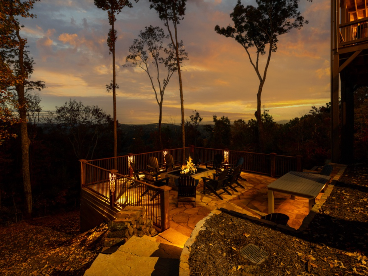 Outdoor patio with a fire pit and seating area overlooking a wooded mountain landscape at sunset, illuminated by warm ambient lighting.