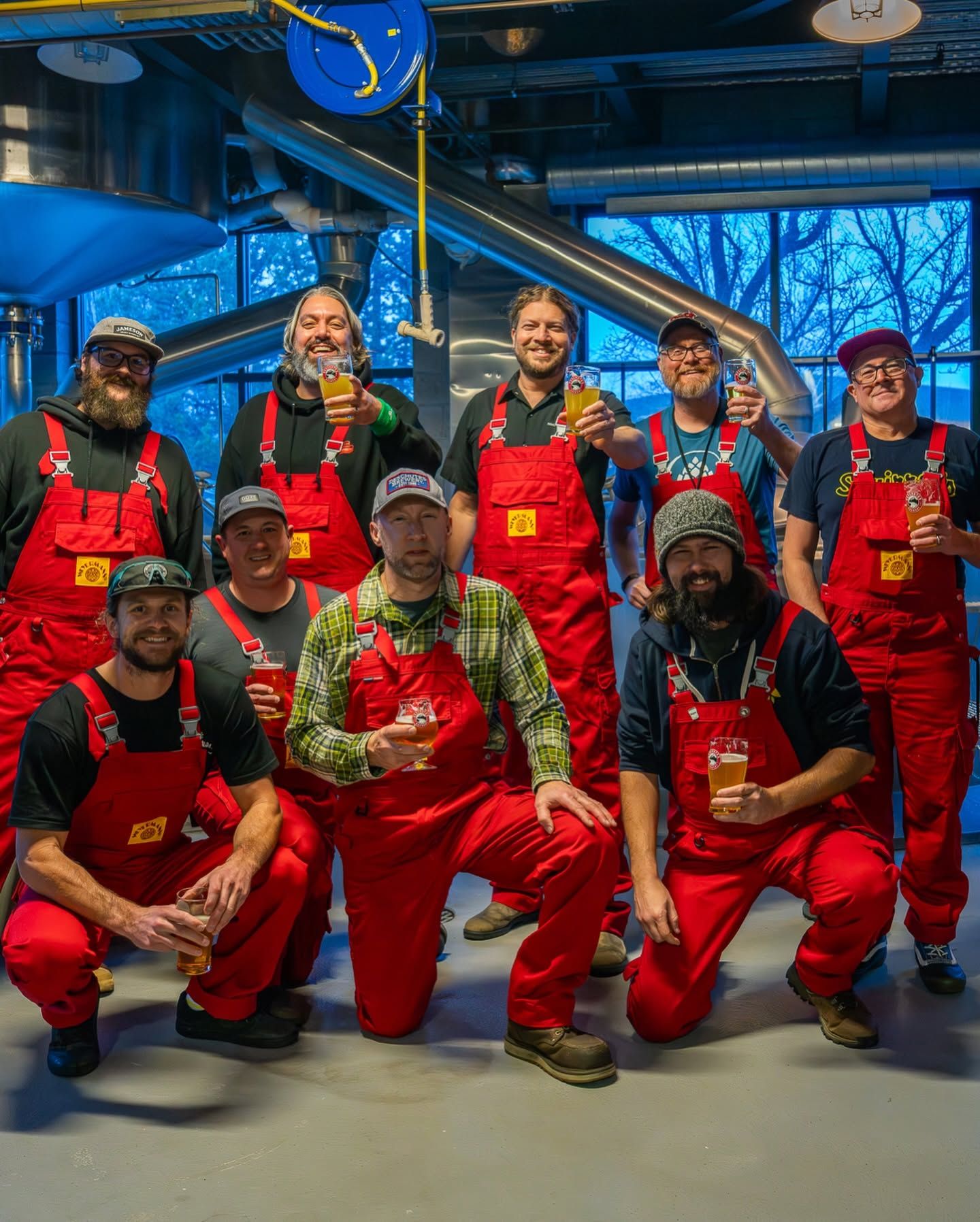 The brewing team at Deschutes Brewery in Bend, Oregon, poses in their signature red overalls inside the production facility, raising glasses of craft beer in a cheerful group toast surrounded by stainless steel brewing equipment. 