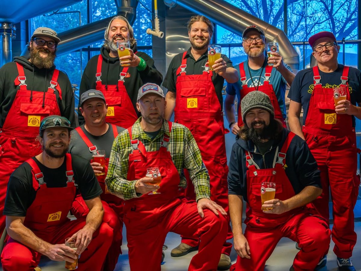 The brewing team at Deschutes Brewery in Bend, Oregon, poses in their signature red overalls inside the production facility, raising glasses of craft beer in a cheerful group toast surrounded by stainless steel brewing equipment.