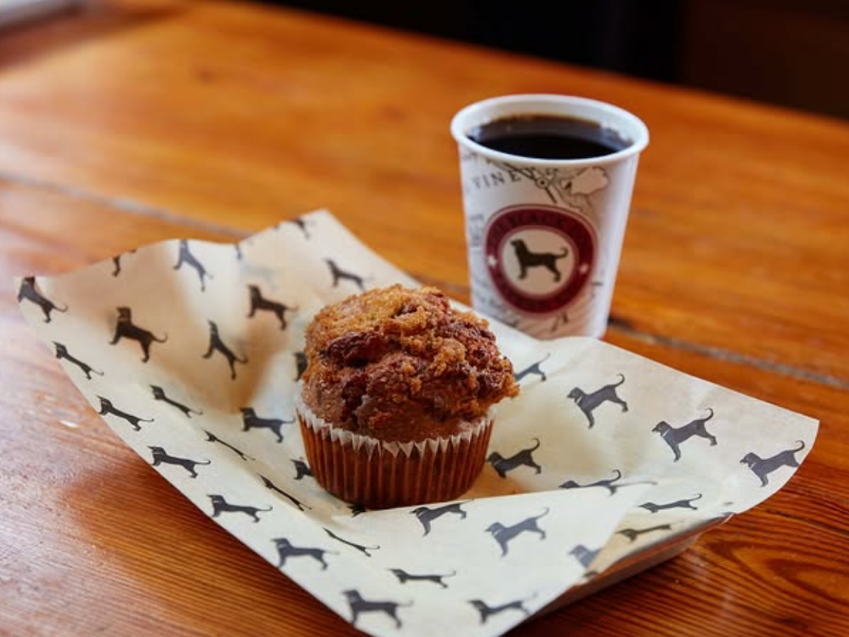 Fresh muffin with a crumb topping served on dog-print parchment paper alongside a cup of black coffee in a to-go cup featuring a dog logo, set on a wooden table.