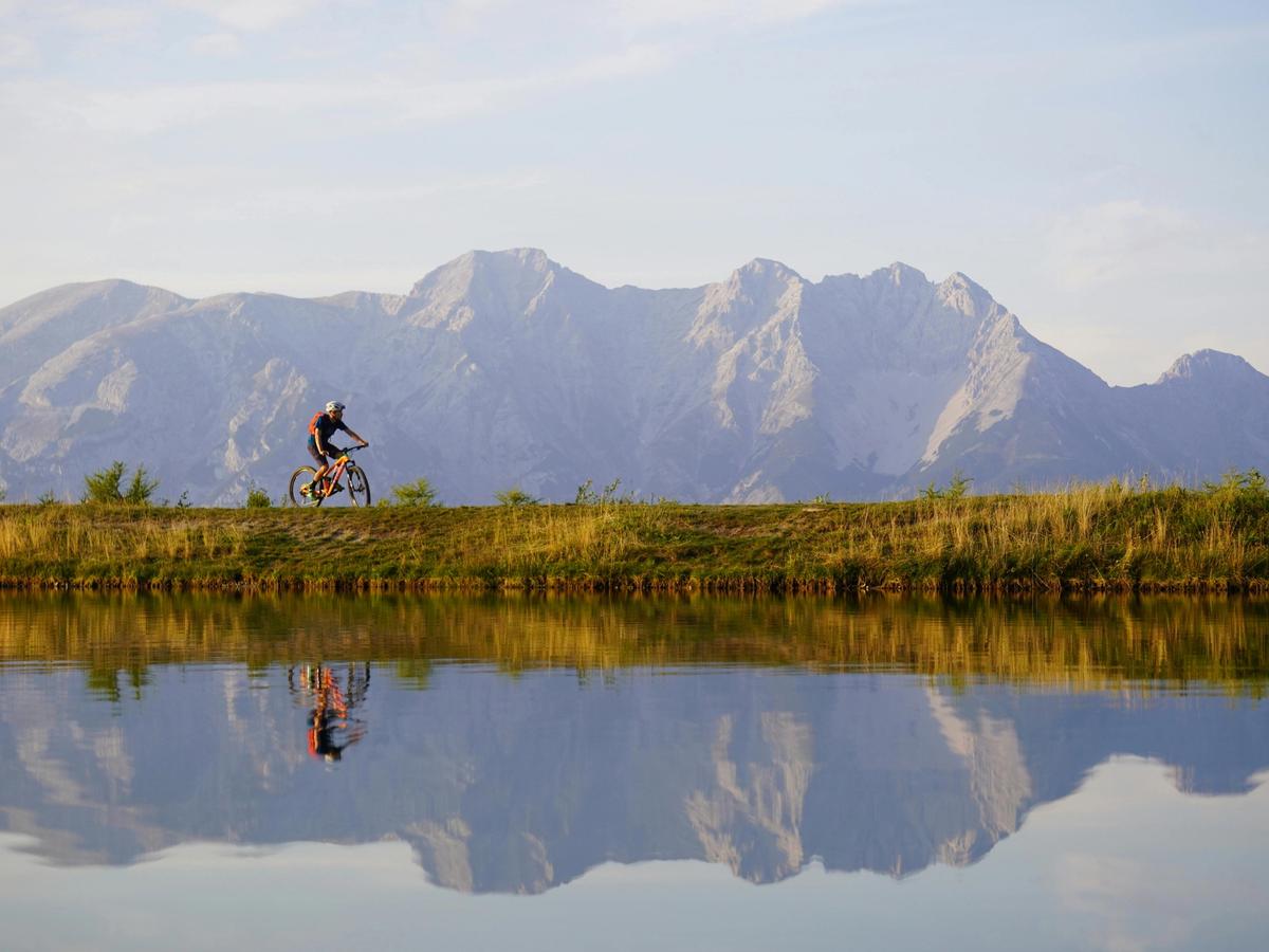A mountain biker rides along a reflective alpine lake with towering peaks in the background. Big Sky’s biking trails offer thrilling rides with breathtaking views.