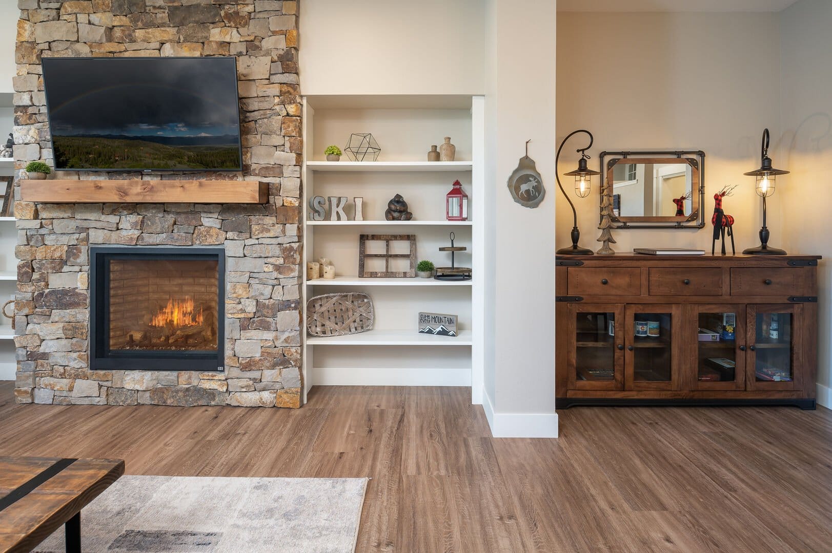 A cozy living room corner with a stone fireplace and mounted TV, flanked by white built-in shelves decorated with rustic and mountain-themed decor, and a wooden sideboard with industrial lamps and a small mirror.
