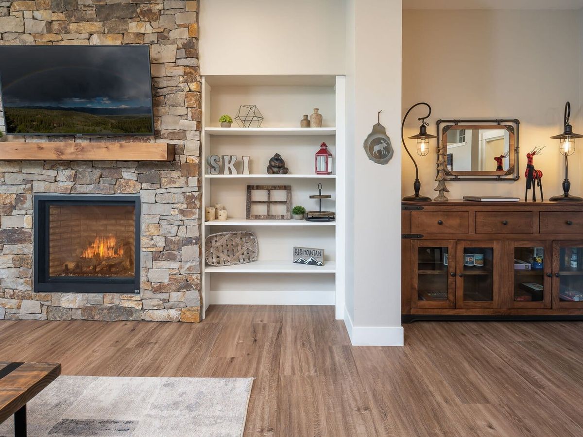 A cozy living room corner with a stone fireplace and mounted TV, flanked by white built-in shelves decorated with rustic and mountain-themed decor, and a wooden sideboard with industrial lamps and a small mirror.