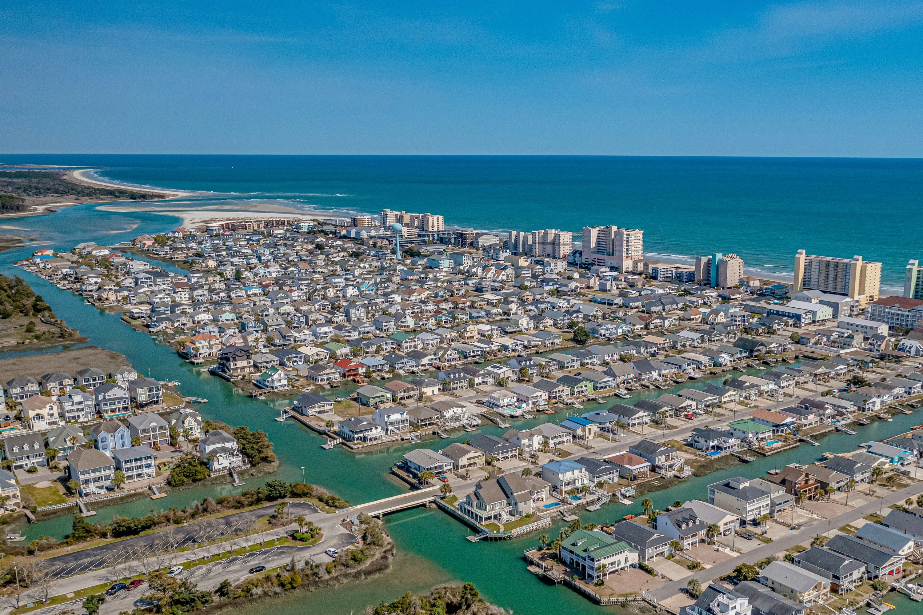 Aerial view of a coastal town with canals and densely packed houses leading to high-rise buildings along the beach and turquoise ocean.