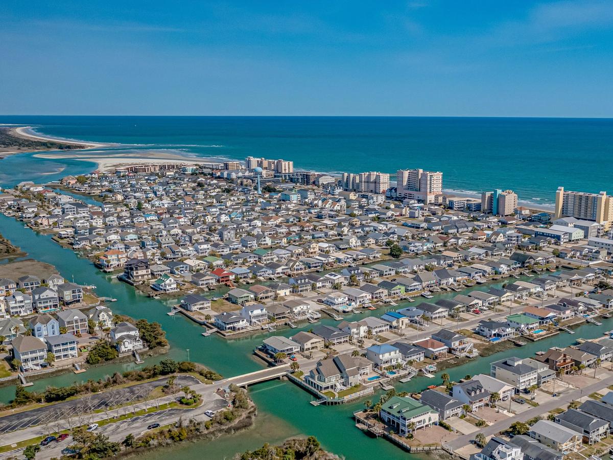 Aerial view of a coastal town with canals and densely packed houses leading to high-rise buildings along the beach and turquoise ocean.